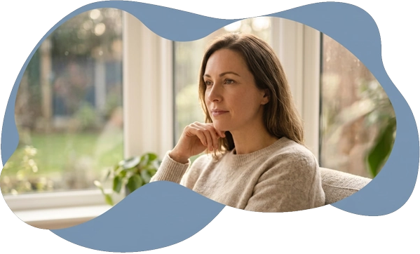 A thoughtful woman looking out a window, framed by a soft blue organic shape, illustrating the search
             for clinical certainty and a foundational roadmap through a formal adult autism assessment.