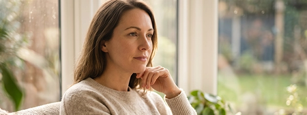 An adult woman reflecting by a window, representing the self-understanding and
                                clinical certainty gained through a formal adult autism assessment.