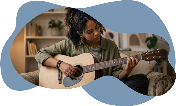 A young woman with curly hair playing an acoustic guitar on a sofa, representing how psychotherapy
             helps individuals navigate neurodivergence, heal from burnout, and live with intention.